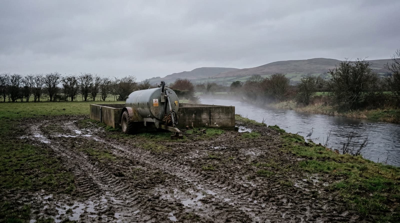 A muddy Welsh farm field at the edge of a rain-swollen river, a slurry tanker parked beside a concrete storage lagoon, low hills in the distance. LIGHT: Overcast late-autumn aftern