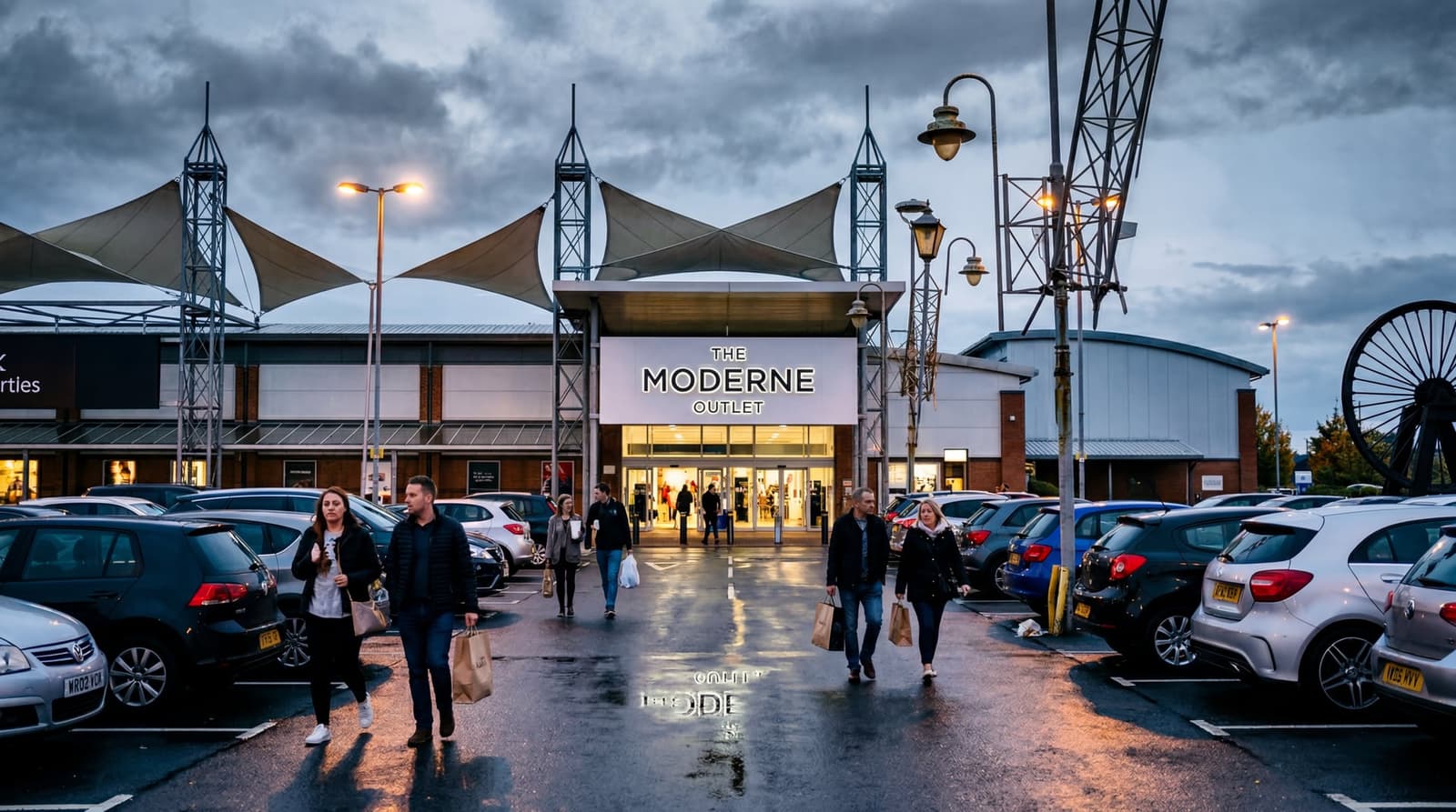 A wide-angle view of a large UK designer outlet shopping centre exterior at dusk, with a freshly installed modern sign reading a retail brand name above the main entrance, cars in